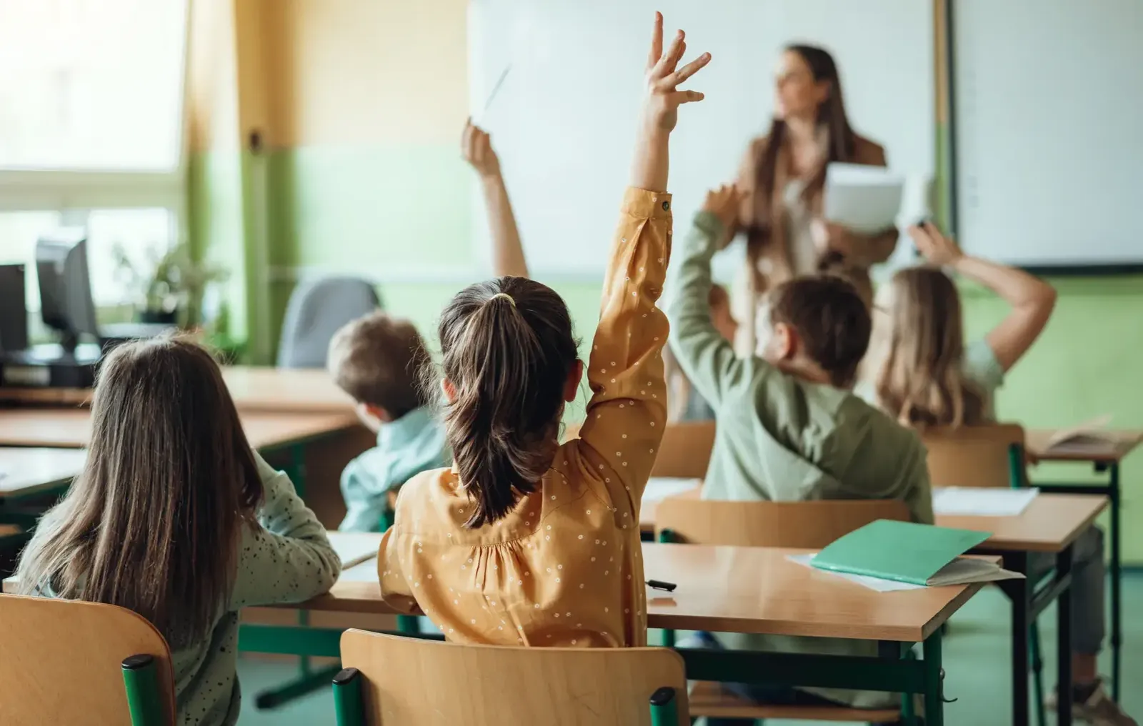 Photo of a class room with a teacher and children, some of them raising their hands.